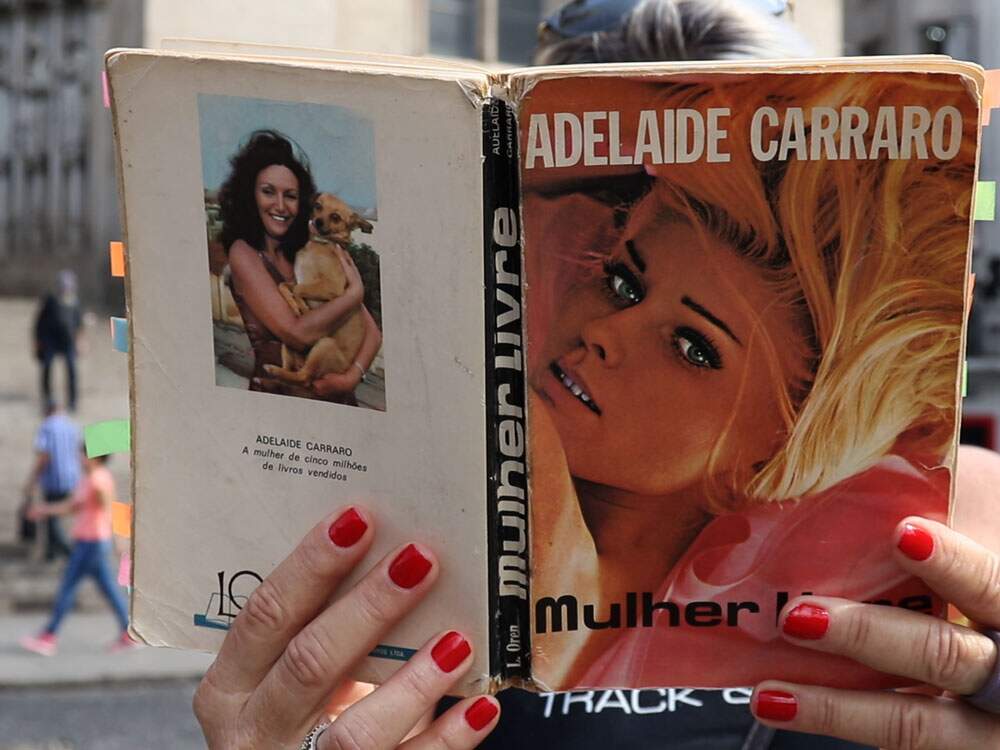A imagem mostra uma mulher com um livro tapando seu rosto. Na capa, uma mulher loira e o nome da autora: Adelaide Carraro. O nome do livro: “Mulher Livre”. A mulher que segura o livro tem as unhas pintadas de vermelho e um anel cinza no dedo médio. Ela mostra parte de seus cabelos lisos e usa óculos escuros na cabeça. Atrás dela, desfocada, há uma