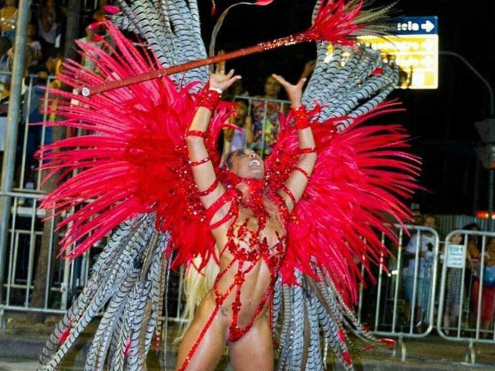 Aline Caldeira está com uma fantasia carnavalesca com predominância do vermelho. Há plumas vermelhas e estampadas em preto e branco. Ela joga um bastão para cima, também vermelho, e se equilibra sobre sandálias com um salto alto.