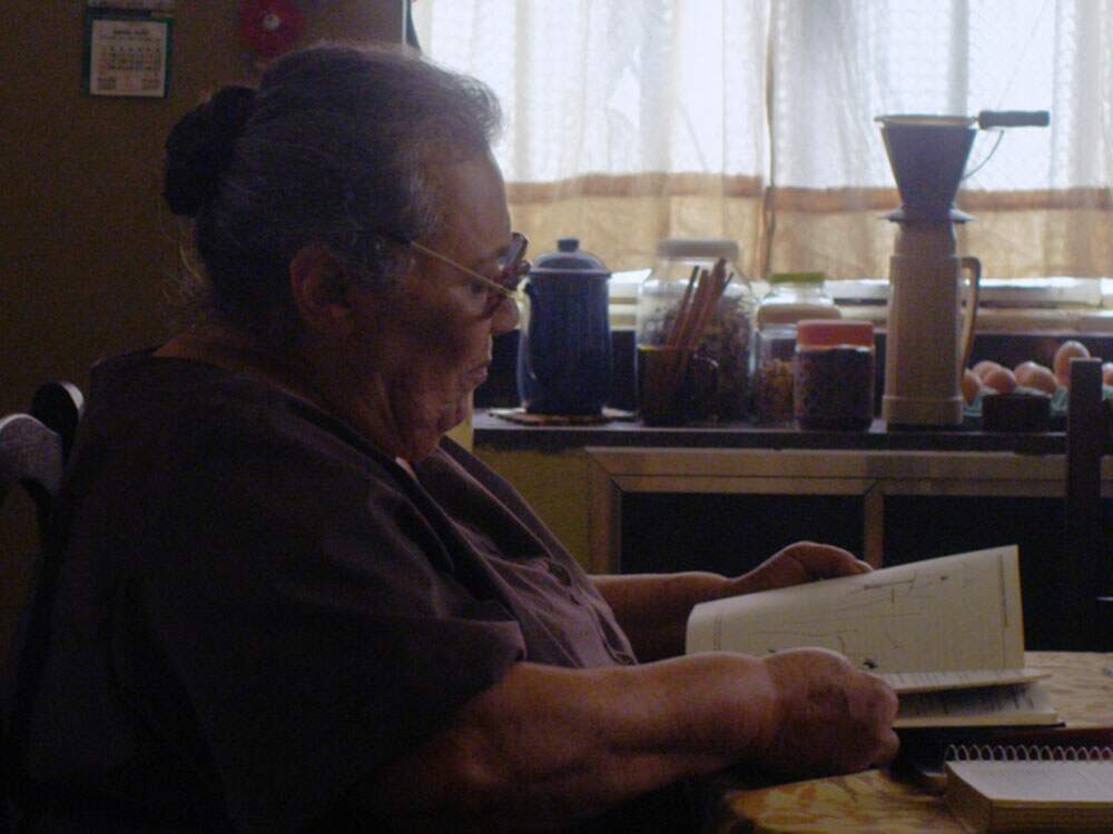 Ângela, vivida pela atriz Teuda Bara, está sentada em uma cadeira junto à mesa de uma cozinha. Ela parece ler anotações em um caderno. Sobre a pia, vasilhas e utensílios se acumulam.