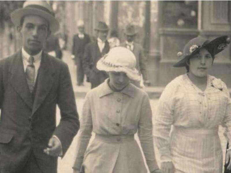 A imagem mostra um homem e duas mulheres caminhando em uma rua, com roupas antigas e chapéus, a foto é preto e branca.