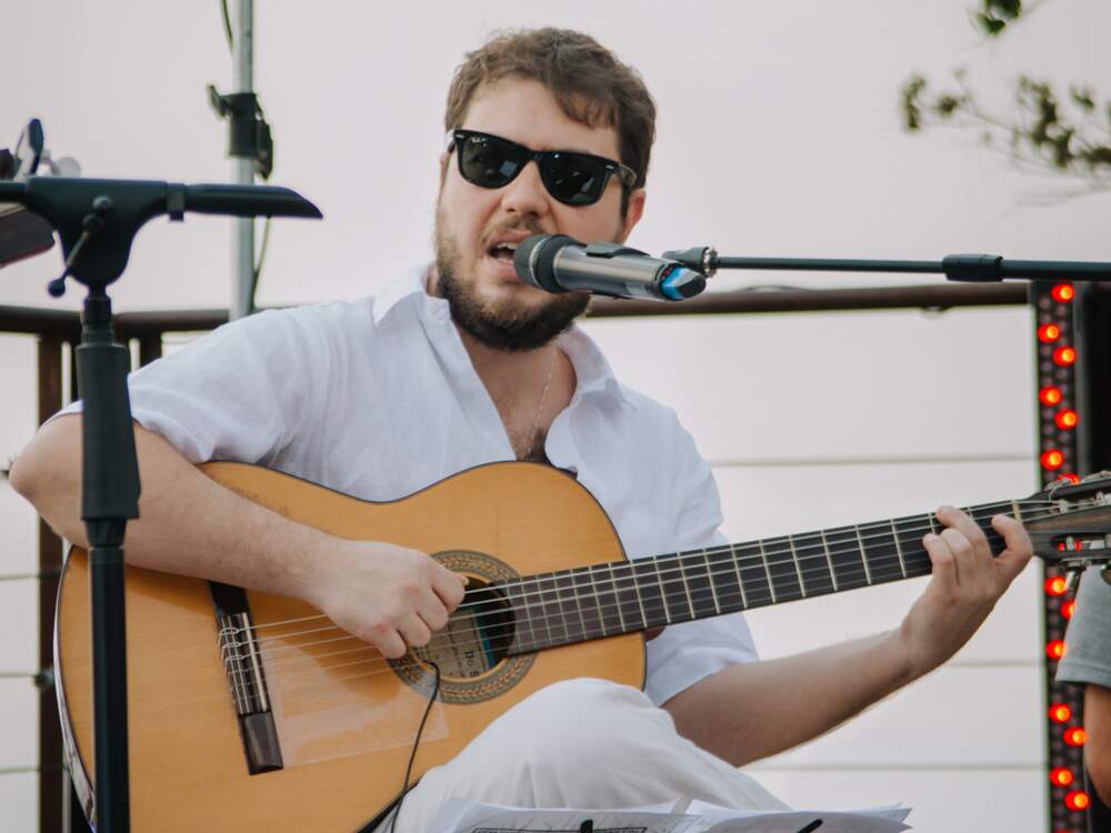 Artur Pádua está assentado, cantando e tocando violão. Branco, tem cabelos e barba castanhos. Usa óculos escuros, camisa branca e calça bege clara.