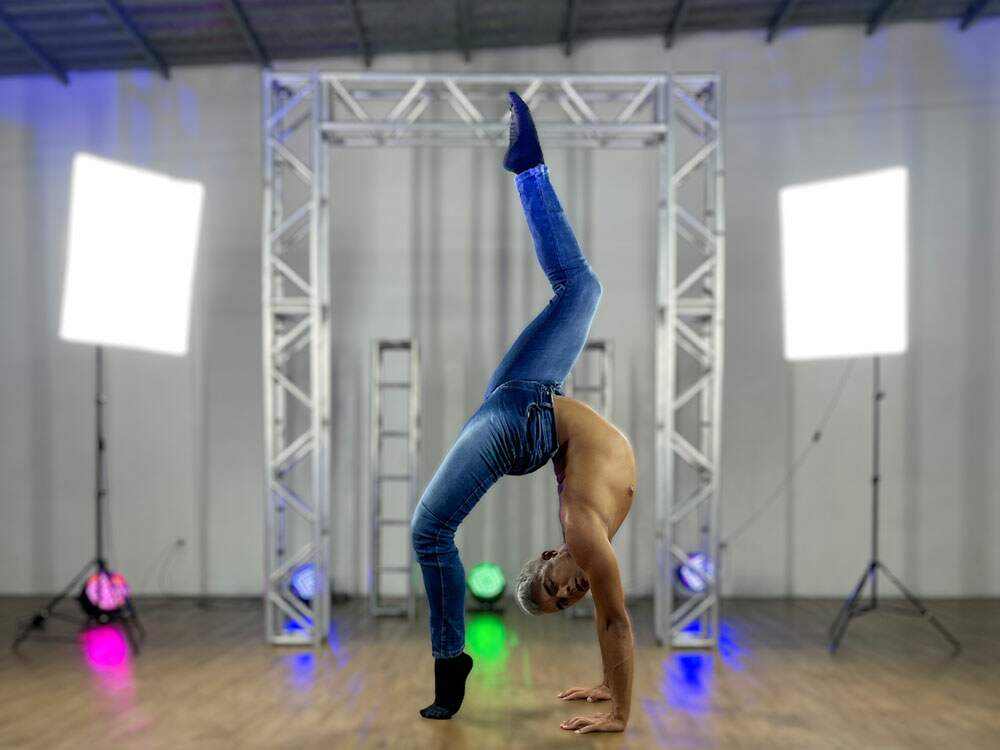 A imagem mostra o artista circense Gabriel Wallace em uma posição de contorcionismo. Com a ponta de um dos pés e as mãos, ele toca o chão e olha compenetrado em direção à câmera. Ele está sem camiseta e calça jeans. Ao fundo, há spots de luzes coloridas.