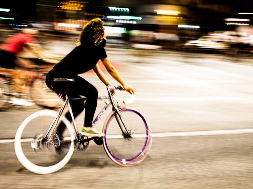 A imagem mostra uma mulher, em primeiro plano, em uma bicicleta fixa. A cidade ao fundo está desfocada, porque ela está em movimento. No entanto, seu corpo da cintura para cima pode ser visto com maior foco. Há luzes ao fundo e uma pessoa em outra bicicleta. Ela usa roupa preta e tem os cabelos encaracolados e tingidos de loiro.