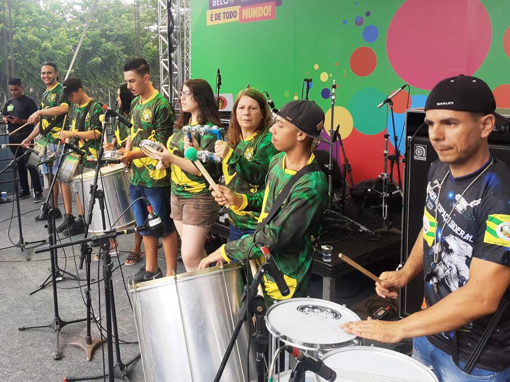 A imagem mostra os integrantes do Bloco Caricato Estivadores do Havaí em um palco. Atrás deles, há uma plotagem com a inscrição “Carnaval de Belo Horizonte é de todo mundo”, com o fundo na cor verde.