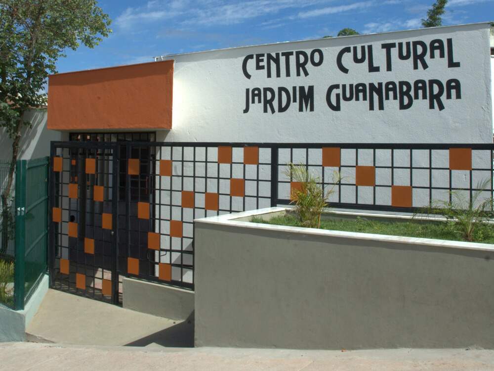 A imagem mostra a fachada do Centro Cultural Jardim Guanabara. Um pequeno jardim está localizado diante de um muro e, acima, está escrito o nome do centro. Há um portão estruturado em quadrados e o acesso é feito por uma rampa.