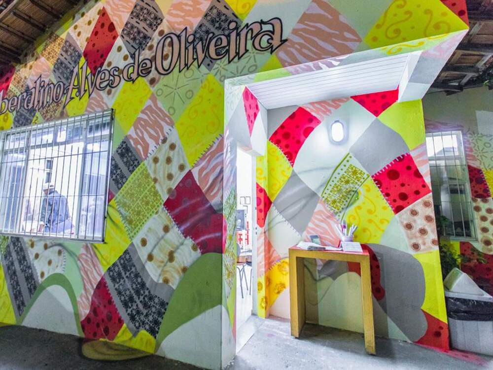 A imagem mostra a fachada do Centro Cultural Liberalino Alves de Oliveira com uma pintura que simula uma colcha de retalhos. Há uma janela aberta e com grade e, junto à porta de entrada, um aparador de madeira com canetas e um caderno.
