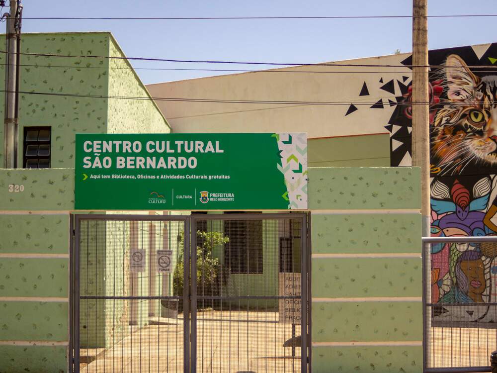 A imagem mostra a fachada do Centro Cultural São Bernardo. Nela podemos ver parte da construção. Ao fundo, há uma parede grande, com um grafitti em diversas cores. A entrada é na cor verde, uma placa centralizada com o nome do centro e uma grade cinza.
