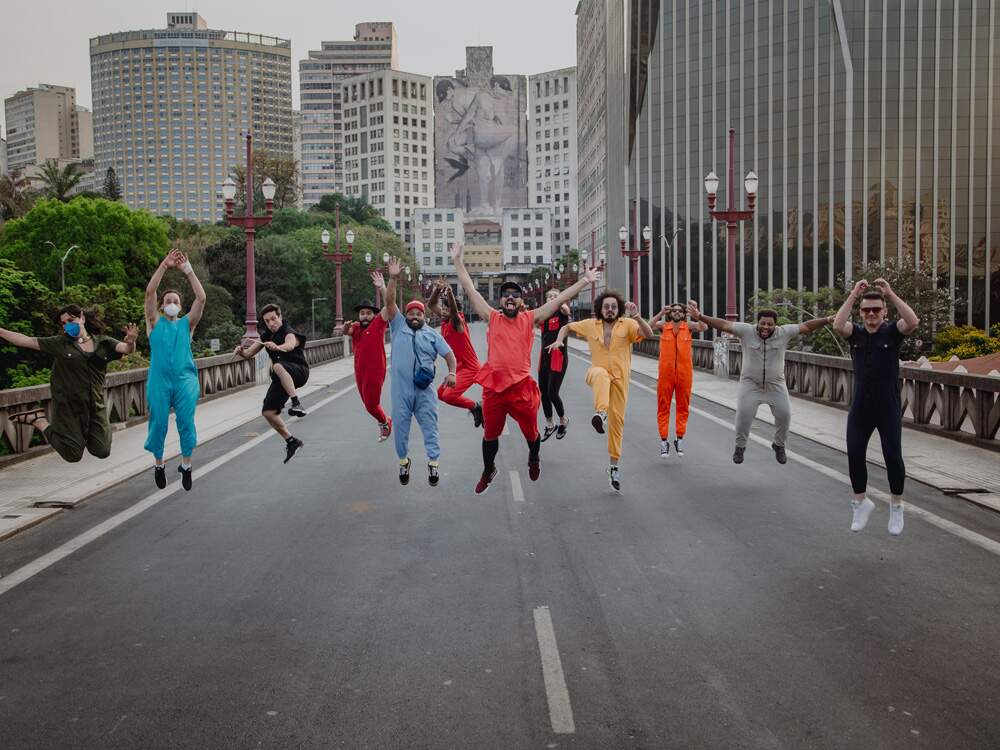 A imagem mostra os integrantes do bloco “Chama o Síndico” no Viaduto Santa Tereza, em Belo Horizonte (MG), com edifícios notórios da cidade ao fundo. A banda foi clicada enquanto pulava. Todos estão usando macacões coloridos e alguns fazem símbolos de coração com as mãos.