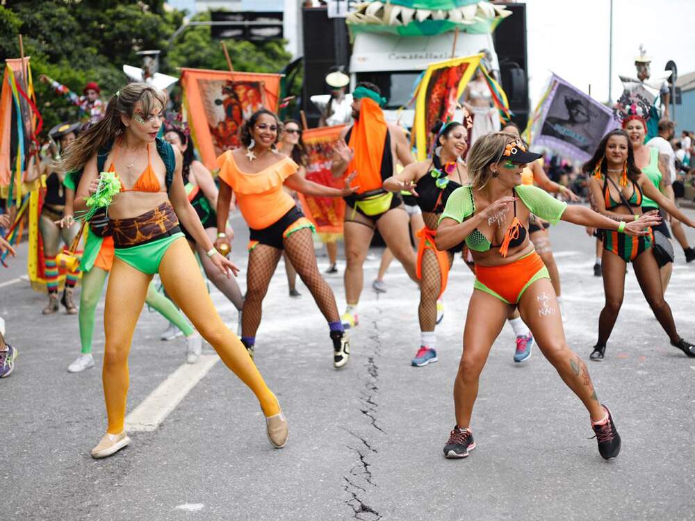 Integrantes da ala de dança do bloco “Chama o Síndico” dançam, tendo ao fundo um trio elétrico. Elas usam roupas alaranjadas e verdes.  