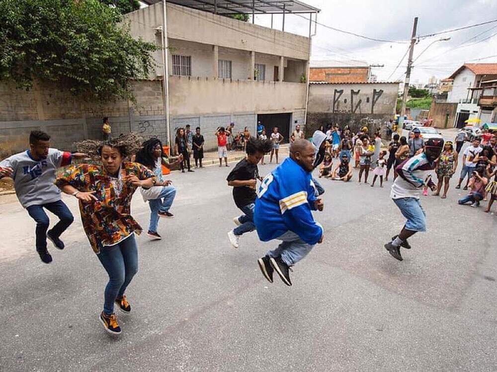 A imagem mostra o grupo “Cia dos Anjos de Danças Urbanas” em uma performance de dança na comunidade. A foto foi tirada no instante em que os dançarinos saltam. No canto direito da imagem, há algumas pessoas da comunidade observando a ação, enquanto outras fotografam.