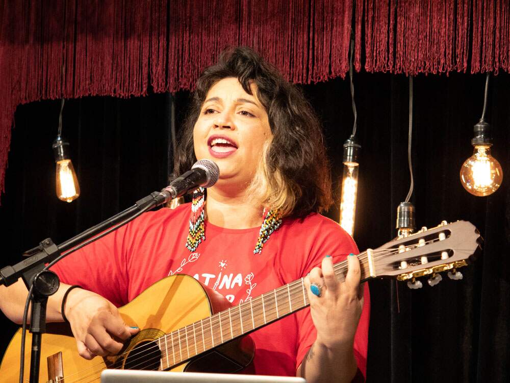 Claudia Manzo usa uma camiseta vermelha e brincos compridos, de miçangas coloridas. Ela toca violão e canta. Seus cabelos são castanhos e ondulados, com uma mecha loira. Ela usa um anel azul, da mesma cor de suas unhas.