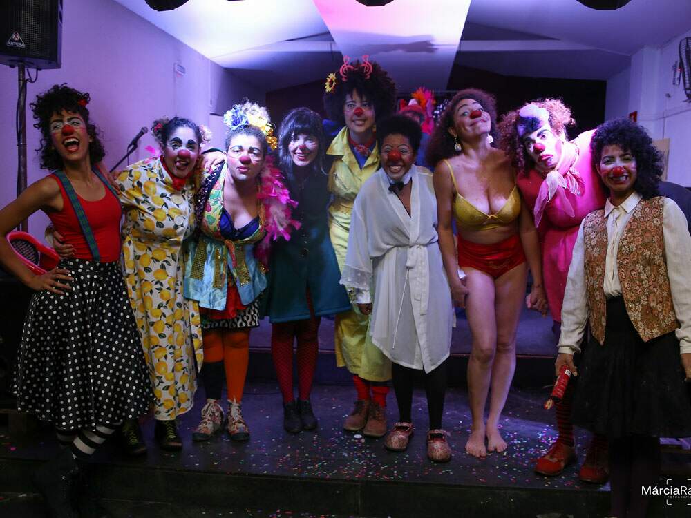 A imagem mostra as integrantes do Coletivo Calcinha de Palhaça em um pequeno palco. São nove mulheres lado a lado, vestidas a caráter, de nariz de palhaça, sorrindo e posando para a foto, com roupas coloridas.