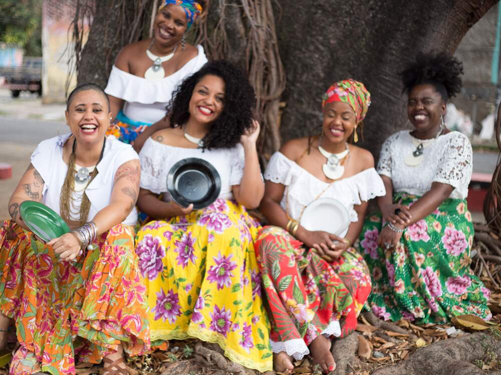 A imagem mostra cinco artistas que fazem parte do coletivo Clã Sambadeiras de Minas. Elas estão sentadas diante de uma árvore de raízes longas. São mulheres pretas e vestem saias de chita e blusas de renda brancas. Todas têm um sorriso largo para a câmera. Três delas seguram pratos fundos coloridos.