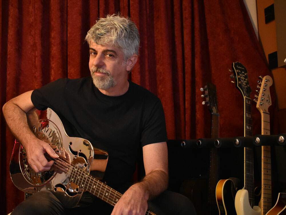 A imagem mostra o artista Rogério Delayon diante de uma cortina de cor terra, ao lado de várias guitarras e violões. Ele está sentado e sorri para a câmera com o rosto de lado, enquanto segura um violão. O músico usa uma calça e camisa pretas e tem cavanhaque e os cabelos grisalhos.
