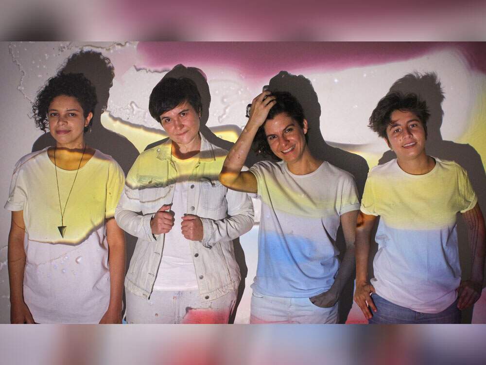 A imagem mostra as quatro integrantes da banda Dolores 602 diante de uma parede. Uma projeção colorida reflete nelas. Elas usam roupas brancas e têm os cabelos curtos. Olham para a câmera sorrindo e com expressão delicada. Em seus corpos e na parede, estão refletidas as cores rosa, amarelo e azul.