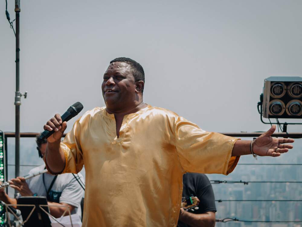 Fabinho do Terreiro canta ao microfone enquanto abre o braço esquerdo. Negro e com cabelos curtos, ele usa uma camisa amarela. Ao fundo, sem foco, prédios da região sul de Belo Horizonte. Na foto aparecem, parcialmente, dois músicos da banda que acompanham o artista.