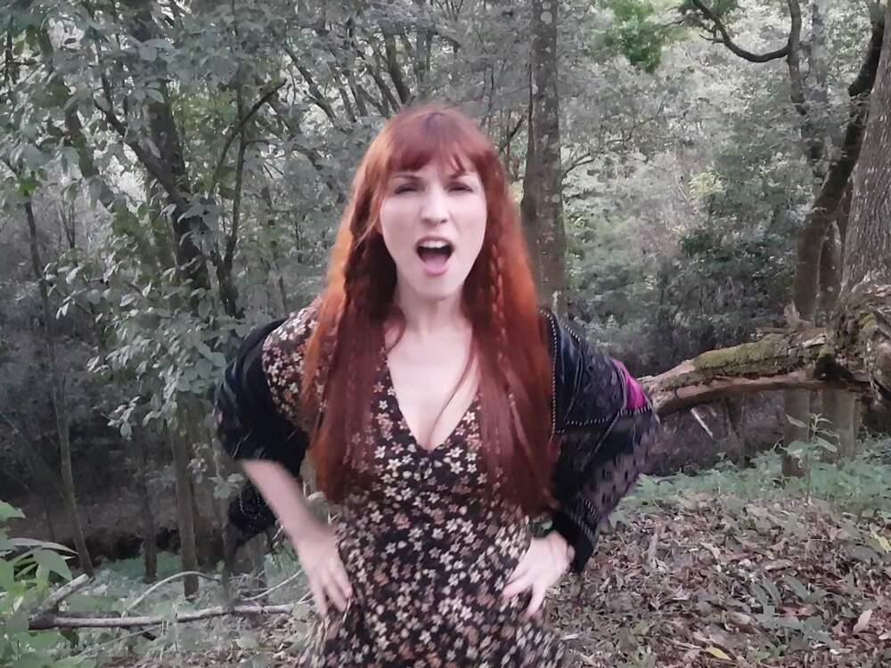 A imagem mostra uma mulher ruiva, de cabelos lisos e compridos, em uma floresta. Ela segura as mãos na cintura e parece dar um grito. Ela usa um vestido escuro com flores e uma jaqueta de couro e renda. 