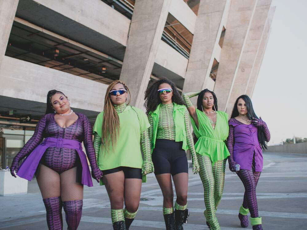 As cinco artistas vestem roupas que variam entre roxo e verde, com detalhes em preto. Uma ao lado da outra, elas olham para a câmera como se caminhassem. Os cabelos variam entre dread, afro e lisos, castanhos e loiro.