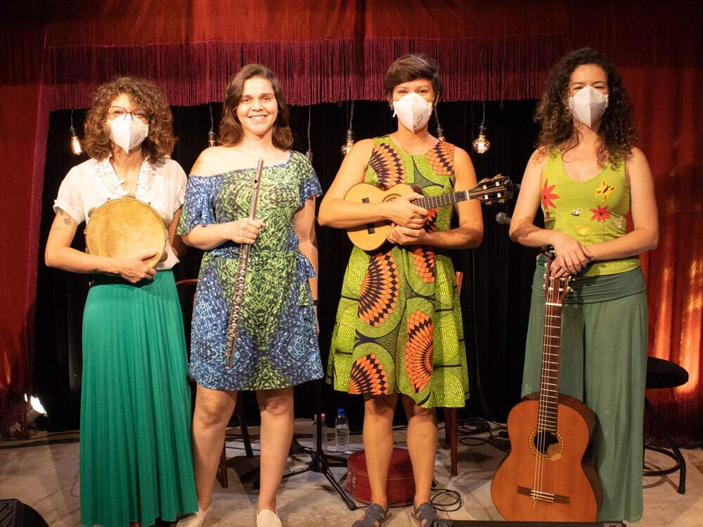 As quatro integrantes do Grupo Chorosas estão em pé em um palco. A primeira à esquerda segura um pandeiro. A segunda está com uma flauta transversal. A terceira artista segura um cavaco. A quarta mulher segura um violão de sete cordas.