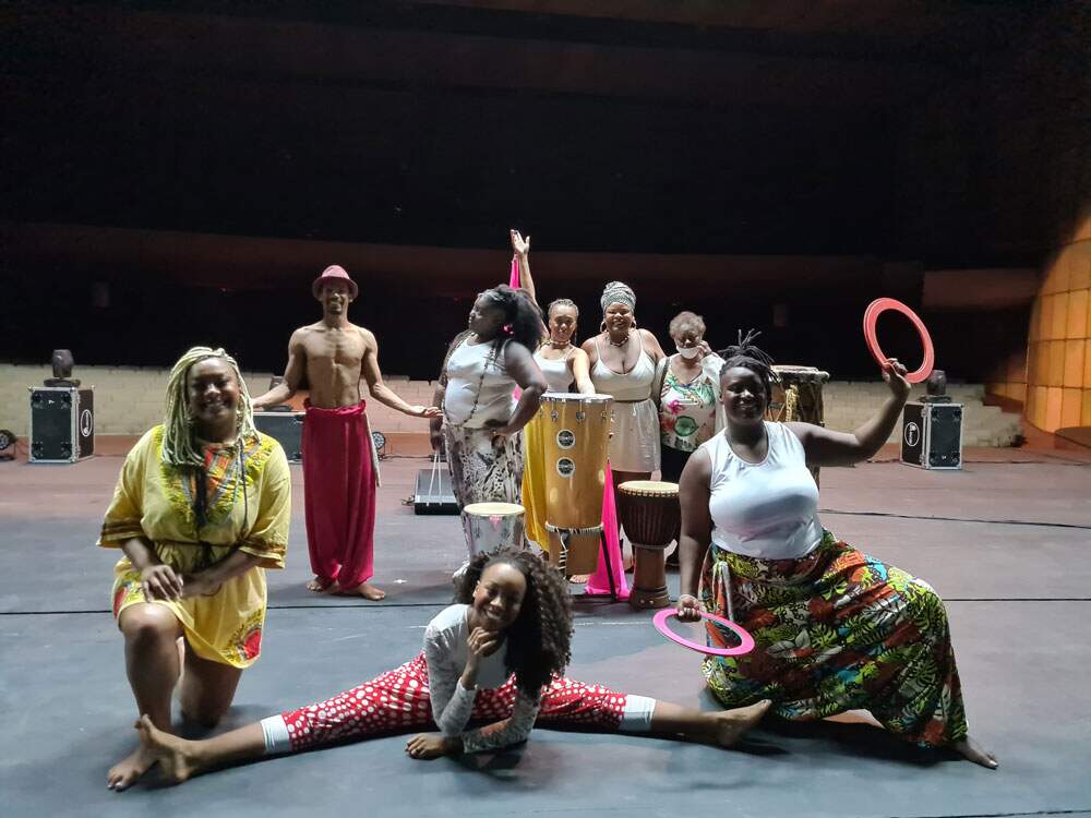 A imagem mostra artistas circenses do grupo “Jotha Produções”. Eles estão em um palco de teatro, de costas para a plateia vazia. No total, há oito pessoas, entre elas sete mulheres e um homem. Todas são negras, usam roupas com tecidos afro, coloridos, e há alguns instrumentos de percussão entre eles.