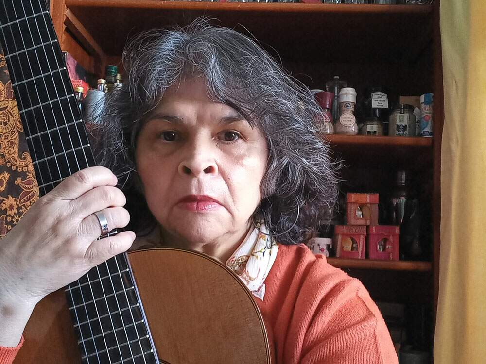 A imagem mostra a cantora Mara do Nascimento ao centro, segurando um violão de pé em seu corpo, e olhando para a câmera. Ela está diante de um móvel com prateleiras e temperos. A artista tem os cabelos grisalhos e usa uma blusa de lã alaranjada.