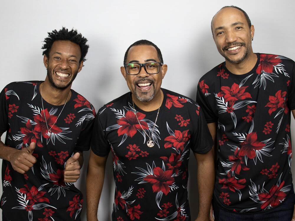 A imagem mostra os integrantes do grupo Black Trio de pé, lado a lado, vestindo uma camiseta de mesma estampa, preta, com flores vermelhas e folhas brancas. Eles posam diante de um fundo branco, sorrindo para a câmera. 