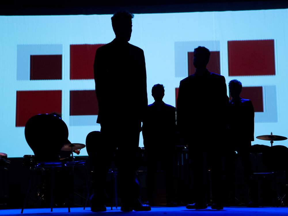 A imagem mostra a silhueta de quatro homens, de costas para a câmera, virados para uma tela onde são projetadas figuras geométricas vermelhas e brancas. Também estão na imagem cadeiras e pratos para bateria.