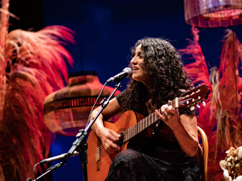 A imagem mostra a cantora Regina Souza sentada em uma cadeira diante de um microfone, segurando um violão e cantando. Ela se encontra em um palco, com um cenário composto de algumas luminárias grandes e plumas coloridas. A cantora é uma mulher branca de cabelos pretos encaracolados e usa uma saia e uma blusa de manga curta.