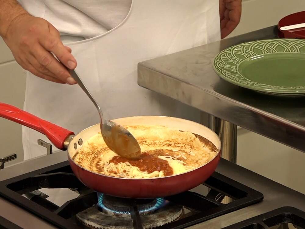 A foto mostra a mão de um homem cozinhando em uma panela, sobre um fogão industrial. Ele usa roupa e avental brancos. Ao lado, uma bancada de inox e, sobre ela, panela, pratos e outros utensílios.