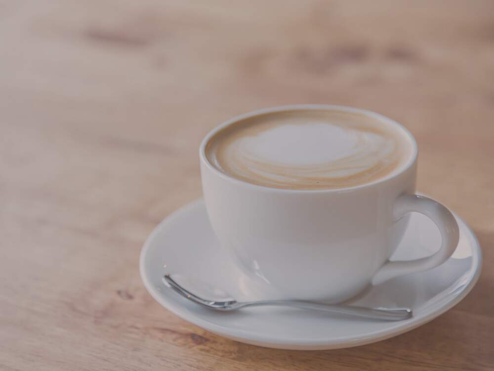 Uma xícara de louça branca, com café com leite e espuma de leite, está sobre uma mesa branca. Há uma pequena colher apoiada sobre o pires.