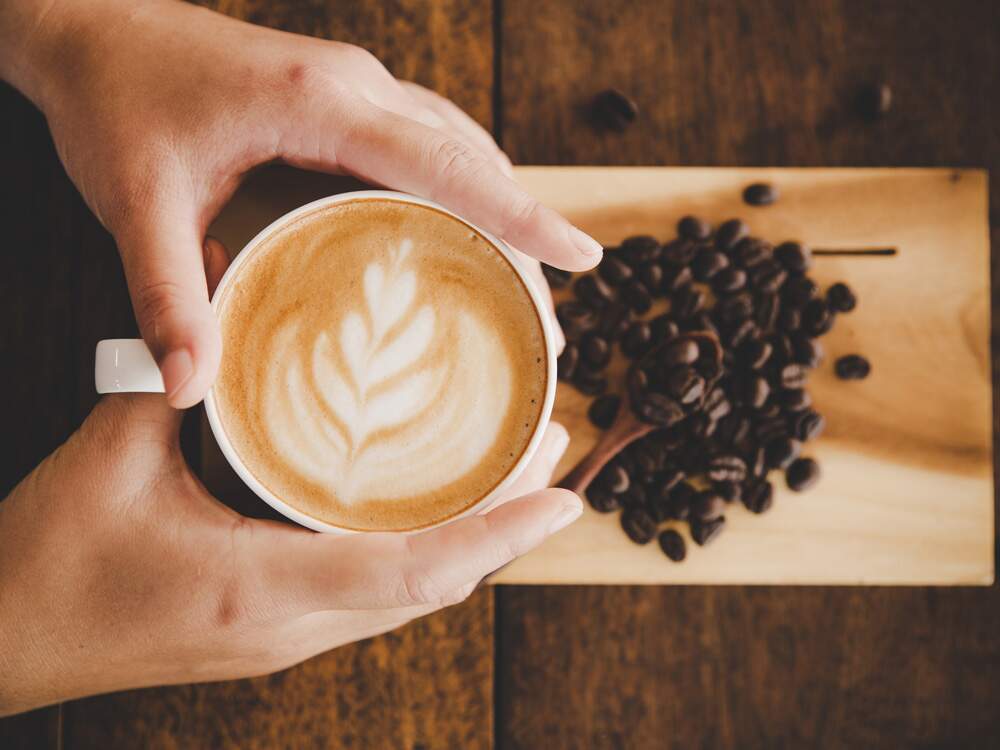 A imagem mostra, de cima para baixo, uma mulher segurando uma xícara de café. Há um desenho de um ramo na espuma da bebida. Abaixo, uma tábua de madeira e grãos de café.