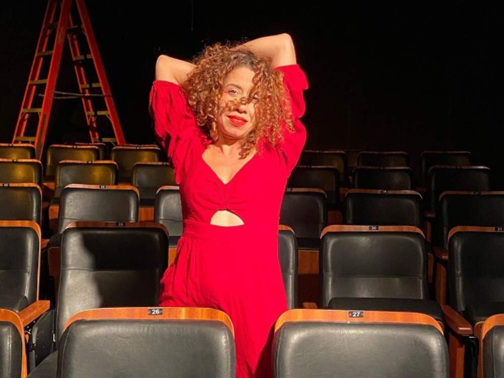 A imagem mostra uma mulher ruiva em meio às cadeiras de um teatro vazio, com os braços cruzados por trás de sua cabeça. Seus cabelos são encaracolados e passam levemente pelo seu rosto. Ela é uma mulher branca, usa um vestido rosa e um batom vermelho. Ao fundo, há uma escada aberta.