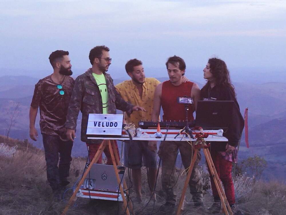 A imagem mostra os cinco integrantes do coletivo de DJs Veludo em um cenário montanhoso, num tom azulado, diante de um set preparado para música eletrônica. Eles estão lado a lado, olhando uns para os outros: da esquerda para a direita, são quatro homens e, no canto direito, uma mulher.