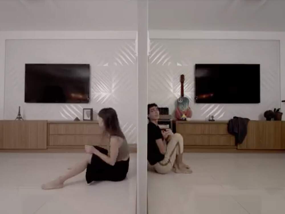 A imagem é dividida em duas partes. À esquerda, uma sala de estar com uma mulher sentada no chão. À direita, a mesma sala, porém de maneira espelhada e com algumas modificações nos adornos. Desta vez, é um homem sentado no chão e encostado na parede que parece dividir os dois ambientes.