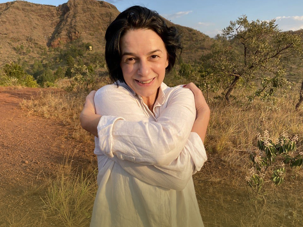 A imagem mostra a bailarina e curadora do Festival Verbo Gentileza 2021, Ana Paula Cançado, diante de uma serra. Ela abraça seu próprio corpo e sorri para a câmera. Ela tem os cabelos pretos e curtos, a pele branca e usa uma camisa branca comprida.
