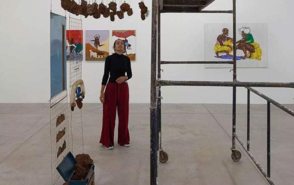 A imagem mostra uma mulher usando calça vermelha, blusa preta em uma galeria de arte. Vários quadros estão pendurados nas paredes