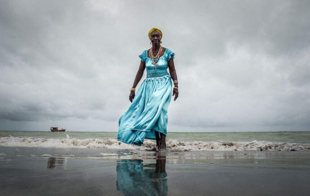 A foto mostra Dona Lia de Itamaracá, uma mulher negra, caminhando pela praia com um vestido azul e turbante amarelo.