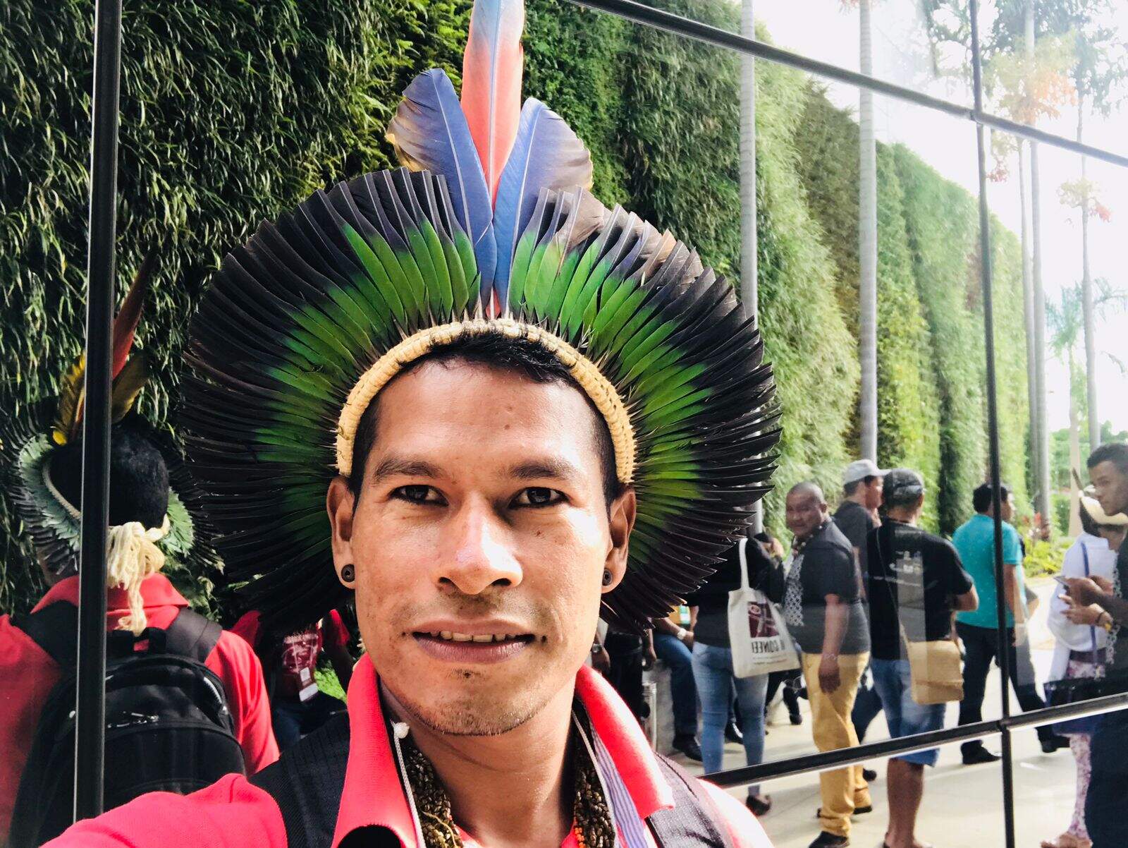 Fotografia colorida de Siwe, homem indígena com cocar na cabeça, ao fundo outras pessoas.