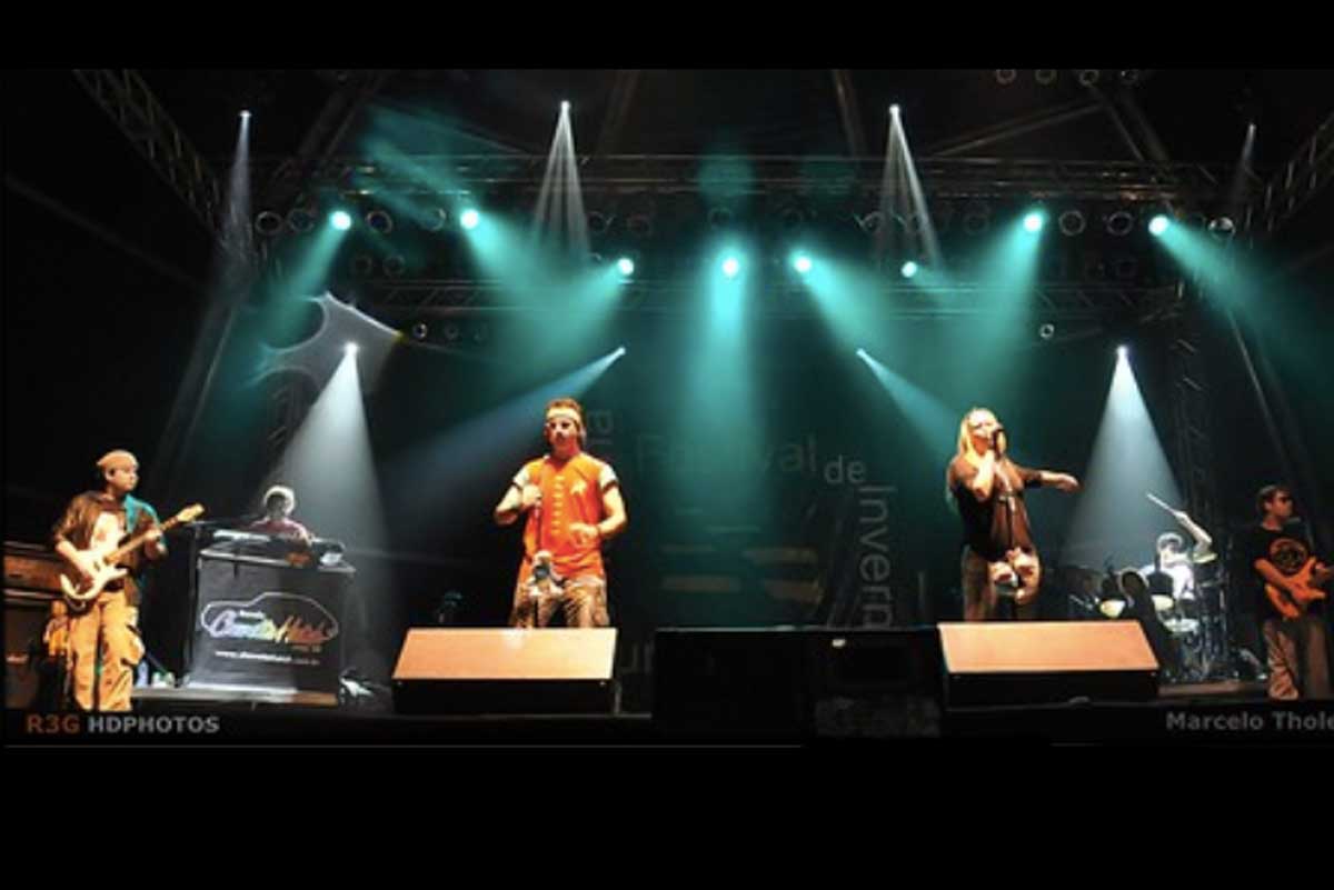 A foto mostra quatro pessoas em um palco de um festival cantando e tocando instrumentos.