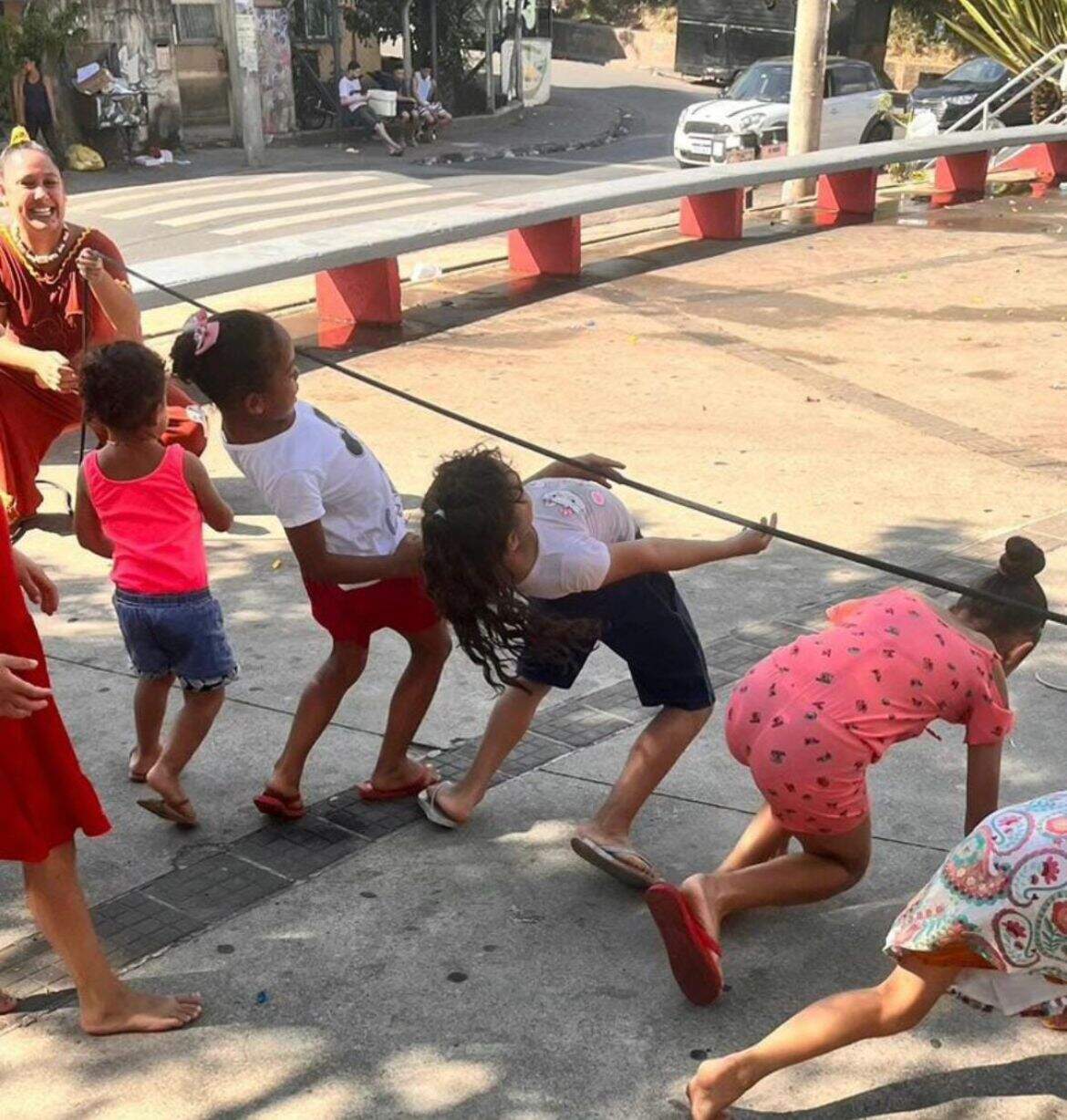 Cinco crianças brincam de passar debaixo da corda, enquanto uma mulher sorridente segura a corda