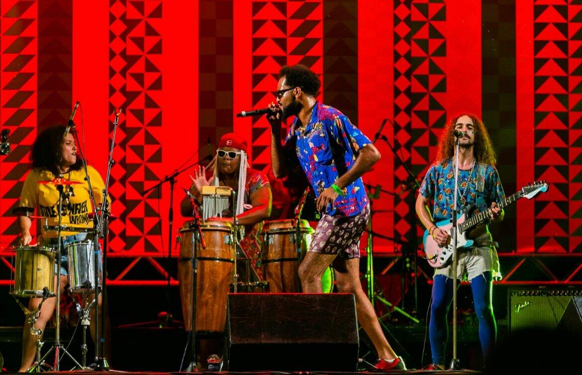 Imagem de um show, com quatro pessoas em um palco. Elas estão com camisetas coloridas. Três pessoas ao fundo tocam instrumentos enquanto um homem com microfone corre para o lado esquerdo 
