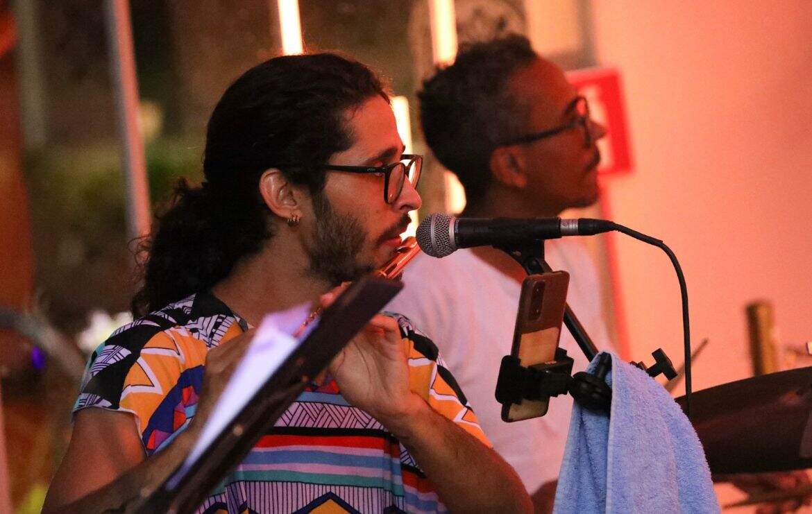 Em primeiro plano, um homem branco de óculos, cabelo comprido amarrado e baba, aparece tocando um instrumento de sopro. Ao fundo há um homem negro de óculos e camiseta branca