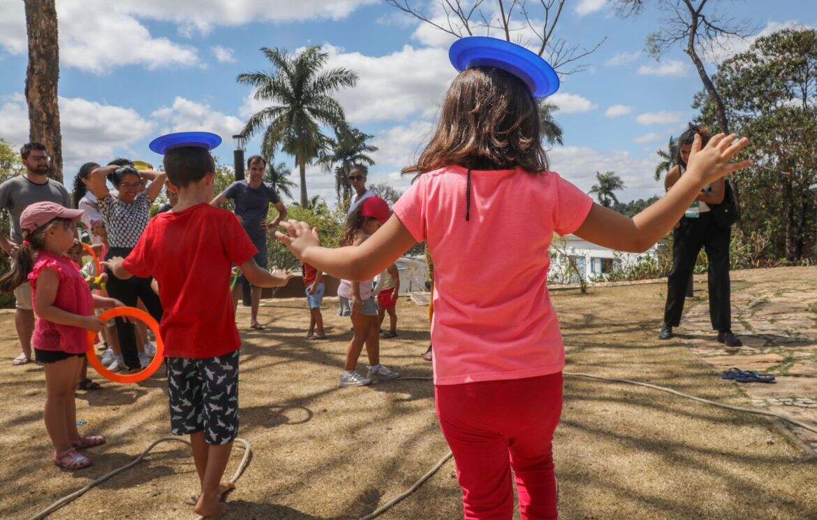 Duas crianças de roupas vermelhas, equilibram um pratinho azul na cabeça. Elas estão em um gramado, Outras crianças aparecem ao fundo
