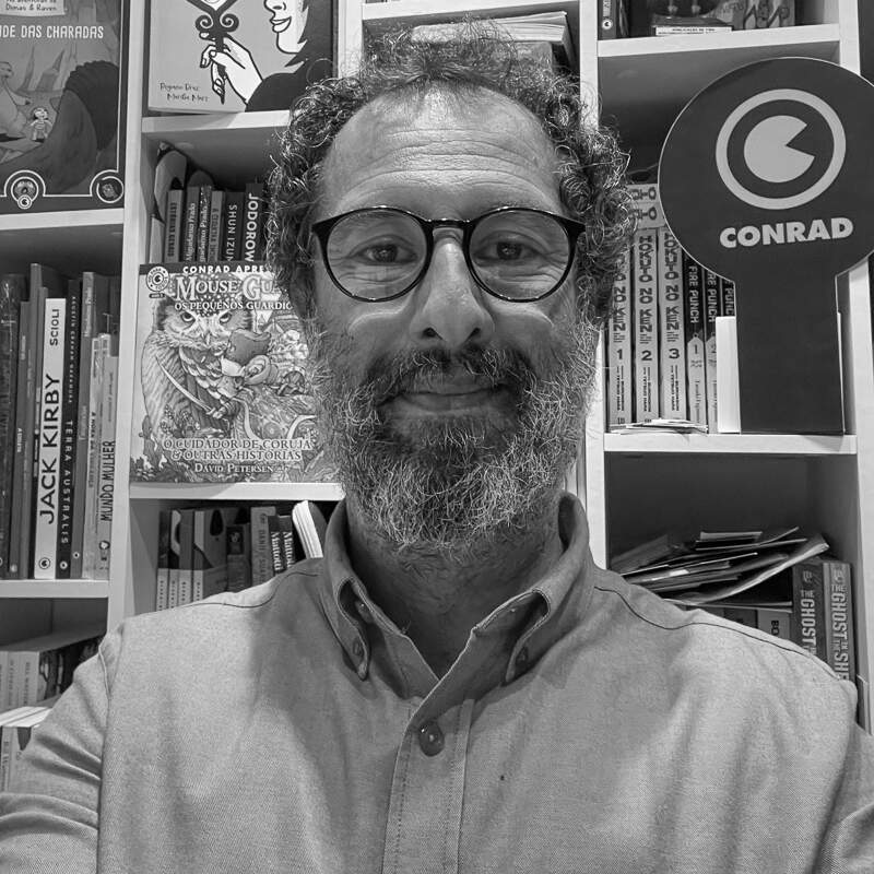  Selfie de um homem de barba em frente a uma estante de livros.