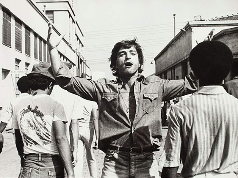 Um homem jovem está de braços abertos e com a camisa parcialmente desabotoada no que parece um pátio de uma fábrica. De costas para ele, trabalhadores caminham, indo em direção contrária,
