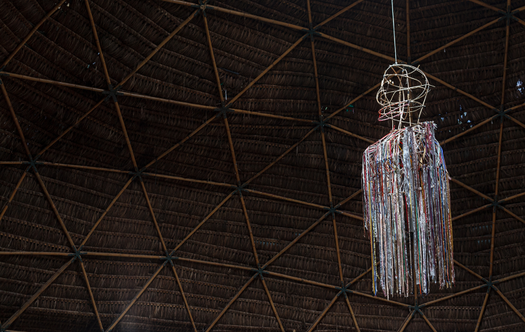 A imagem mostra a instalação Cerbambu Ravena, que é uma obra no formato de um corpo composta de fitas brilhantes numa estrutura, aparentemente de arame, suspensa no teto que esta escuro e é de estrutura de bambu. 