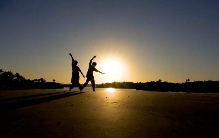 Imagem mostra a silhueta de dois bailarinos dançando na cobertura da casa do baile, em contra luz o por do sol na Pampulha