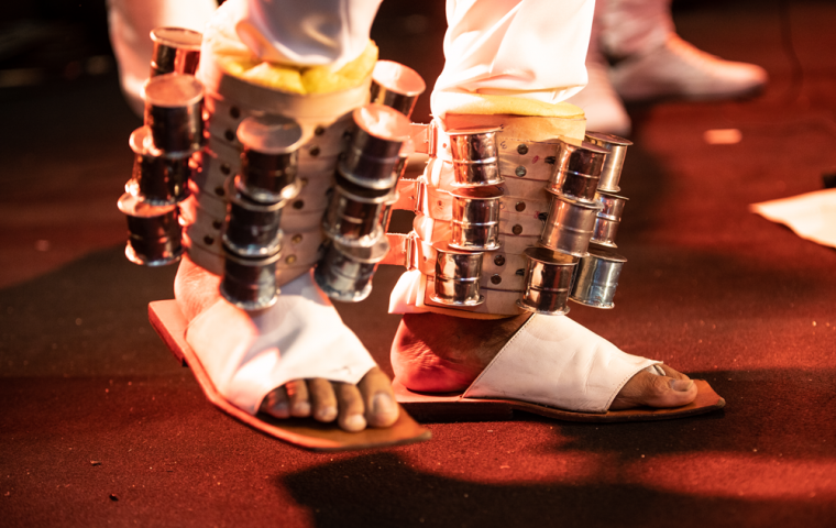 A imagem mostra dois pés com sandálias brancas com instrumentos percursivos presos as eles.