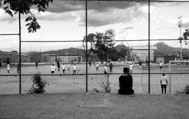 Imagem em preto e branco, com mulher sentada em primeiro plano observando um jogo de futebol em um campo de várzea