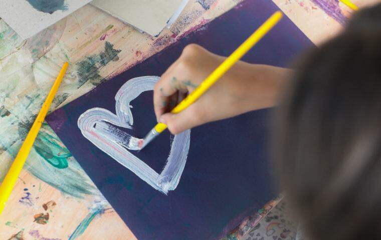 Imagem mostra uma criança de costas no canto da imagem pintando em um papel preto. O foco da fotografia esta principalmente na pintura da menina. É um coração branco em uma folha preta.